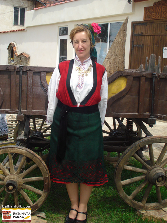 Автентична женска носия от Вакарел Сукман без ръкави с плисе (червена фустанела), риза с бордюри бродерия, зелена вълнена престилка (фута) гарнирана с бордюр кене (самоковска шиена дантела) черна кадифена лента за прикрепване на престилката (кондаци), зел